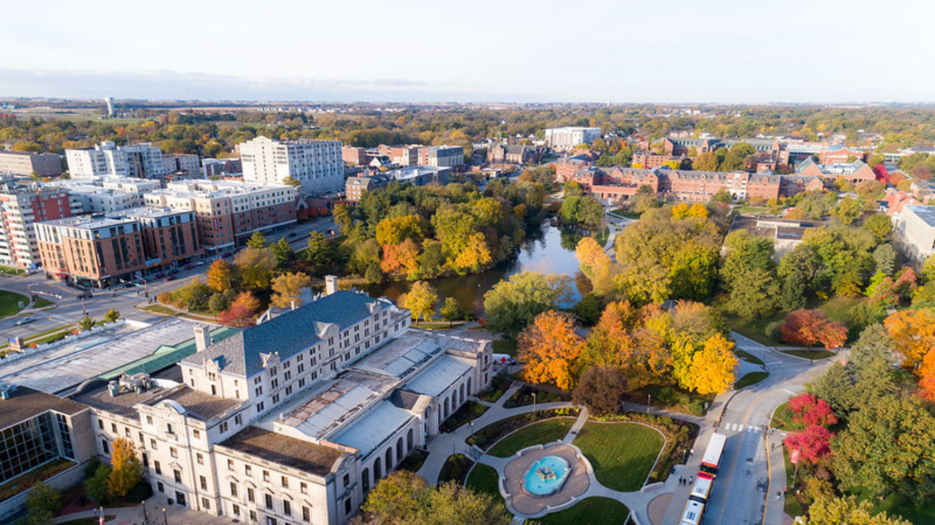 Aerial view of a campus with a large building, fountain, trees in fall colors, and surrounding city buildings.