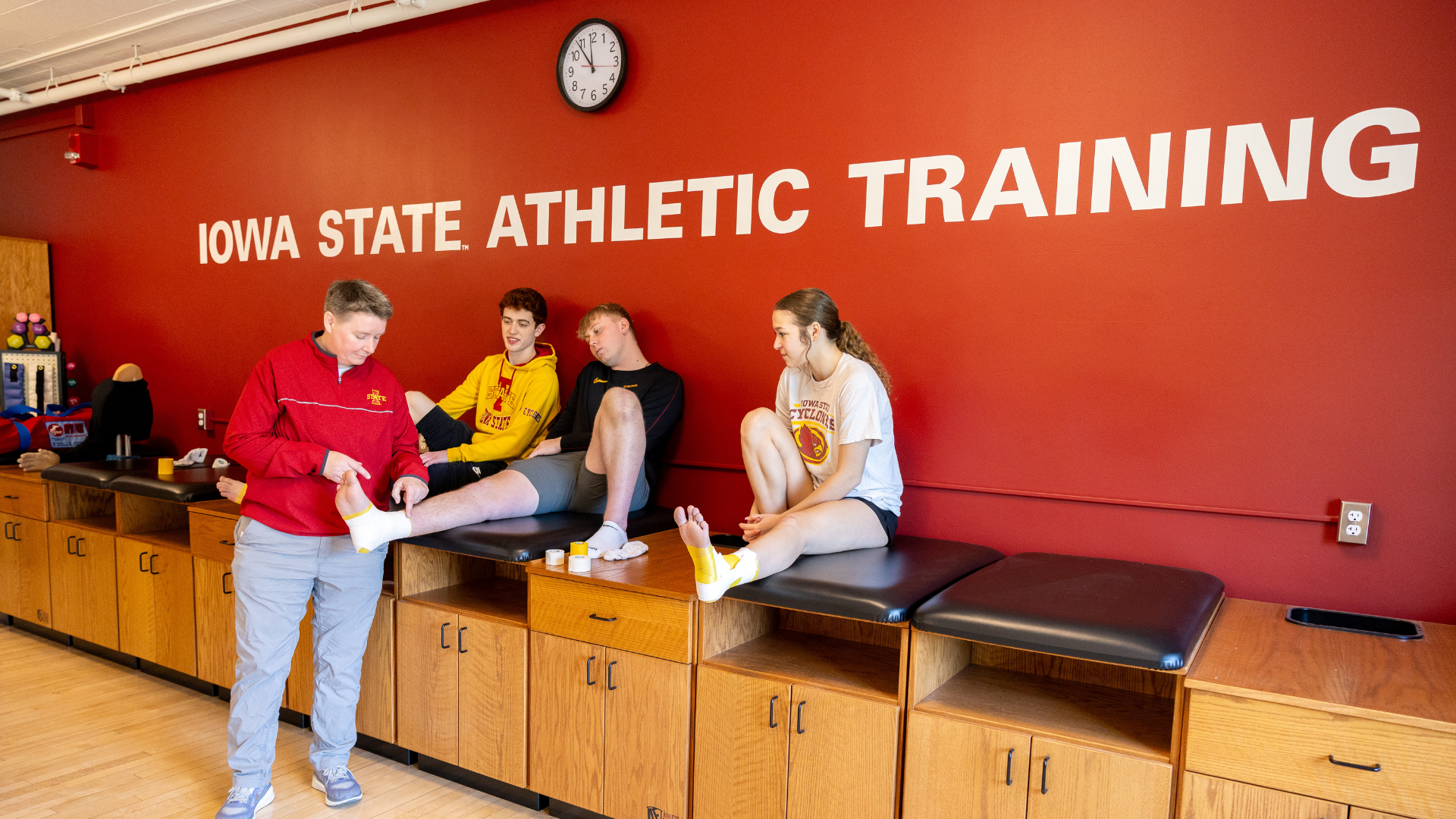 Person taping another person’s ankle while two others sit on treatment tables in an athletic training room.